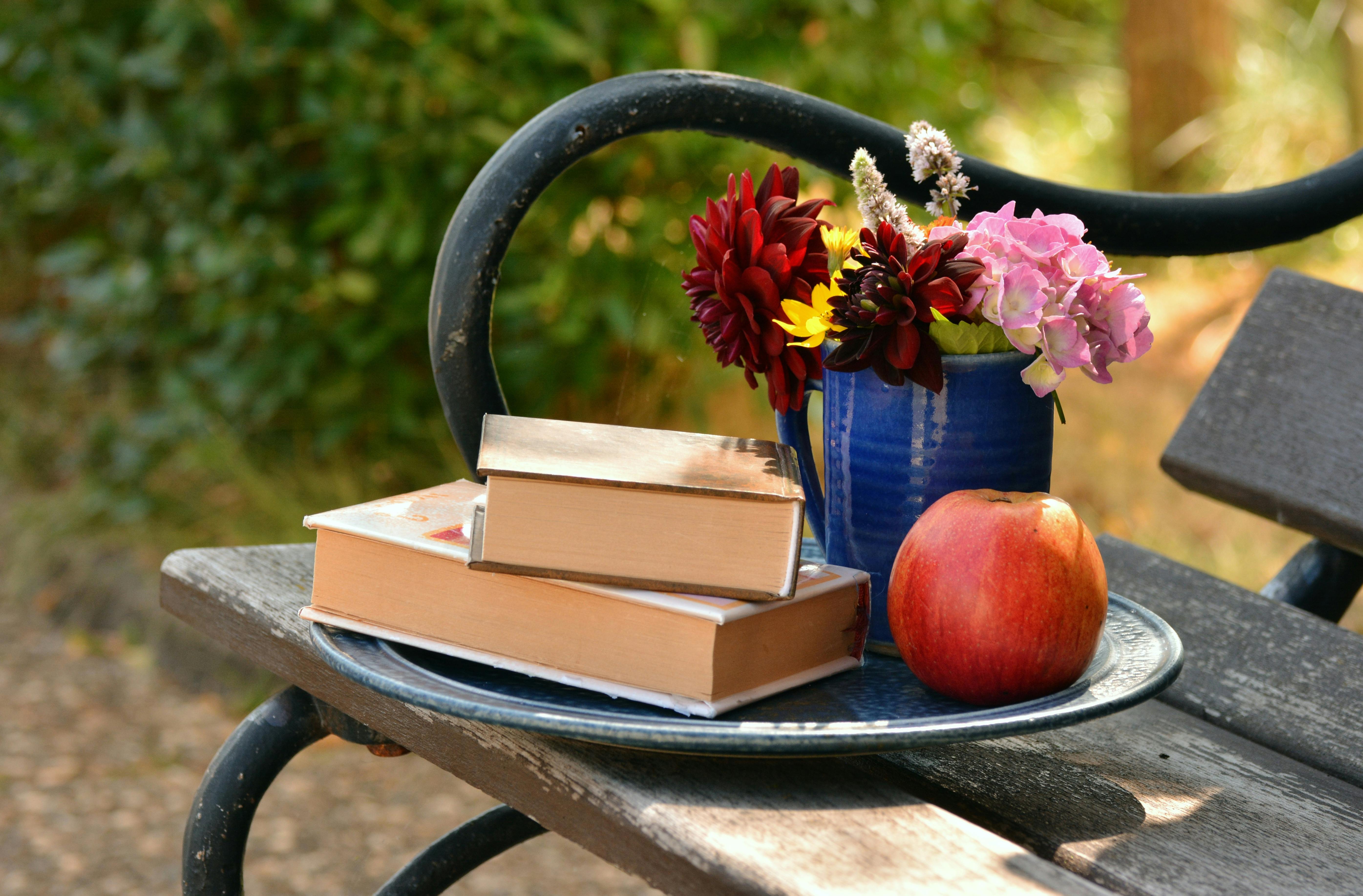 Bench with a plate on it that holds two books, an apple, and flowers in a mug