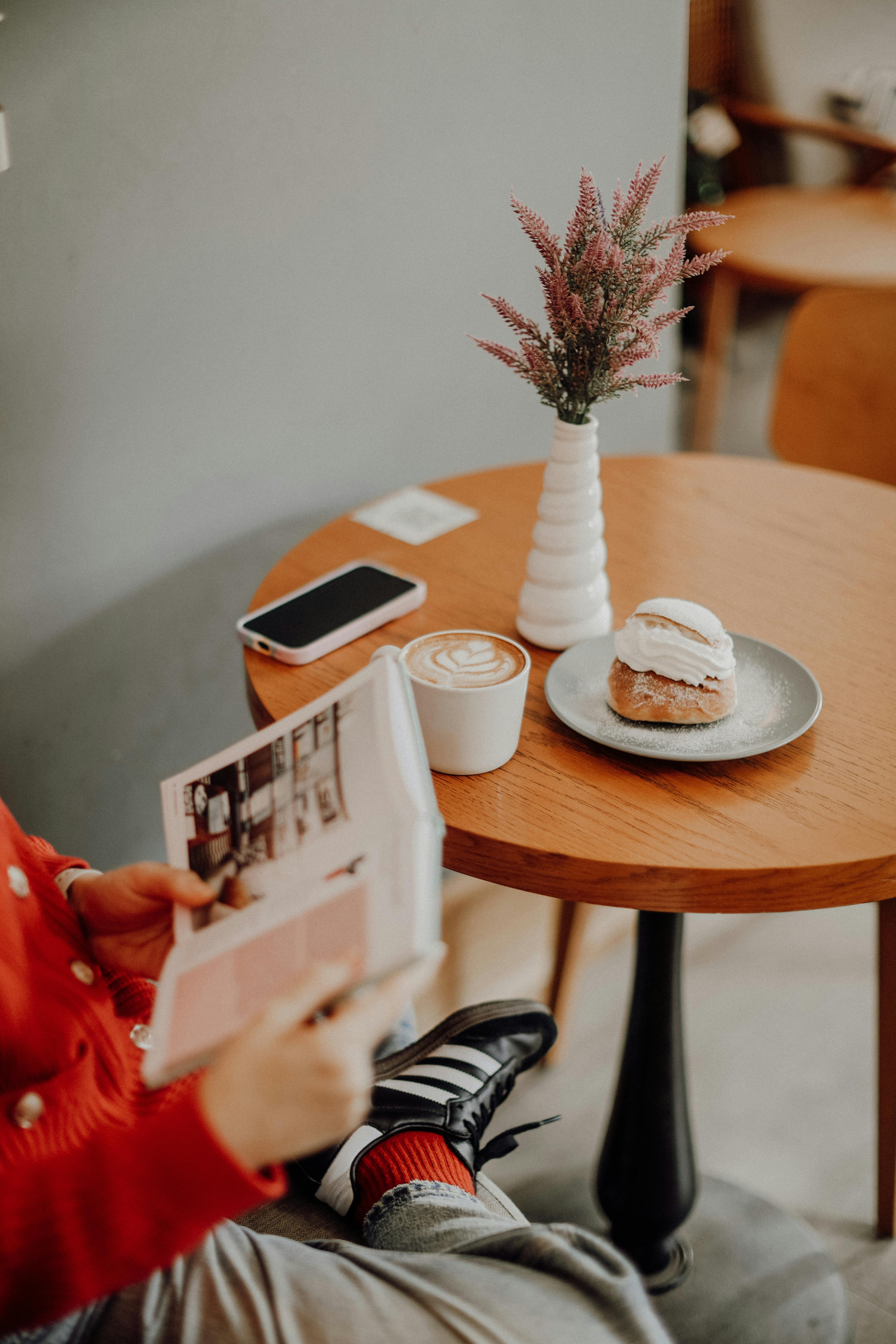 Open book, table with a phone, flower vase with flowers, dessert on a plate, latte in a mug.