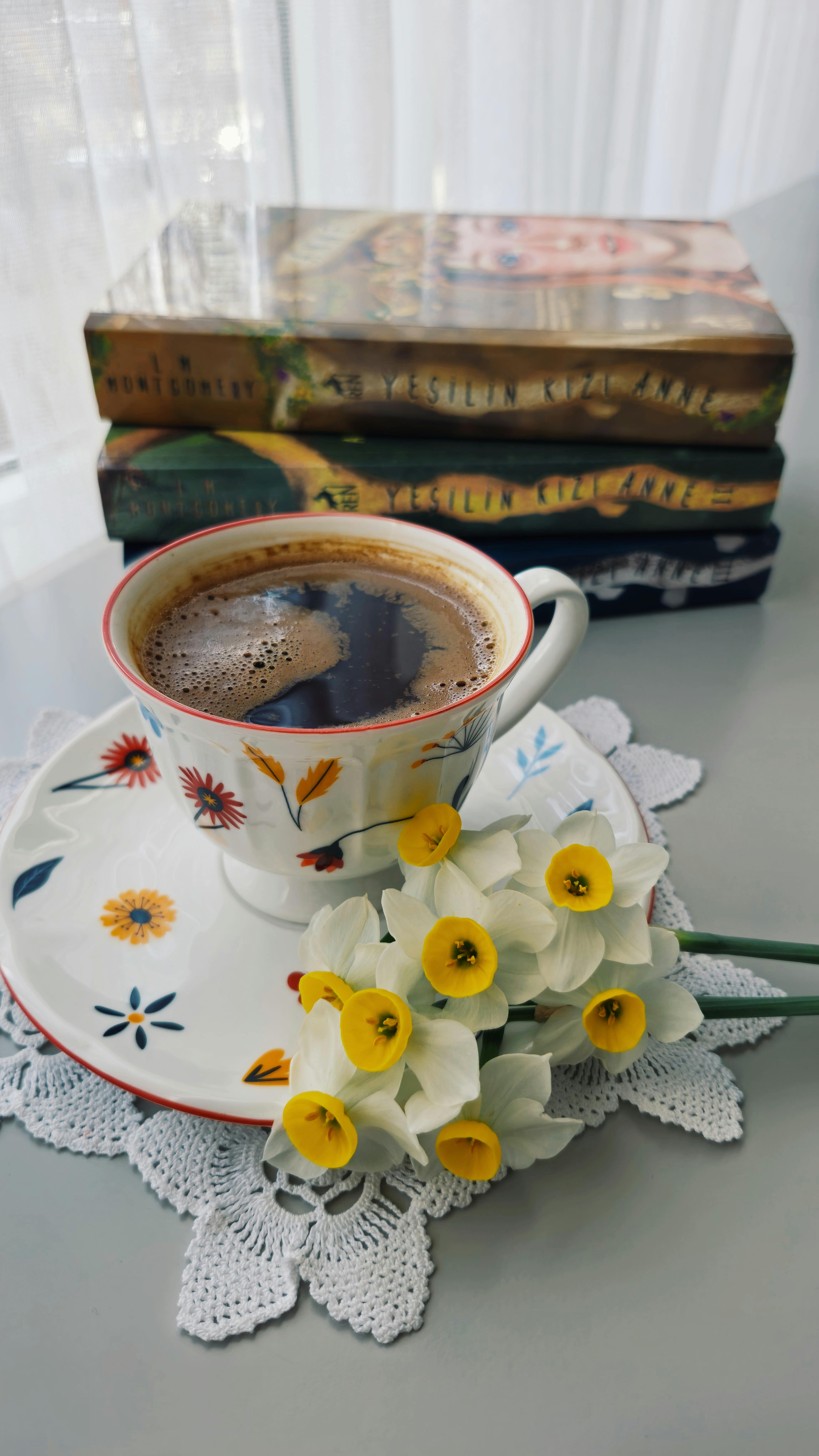 Coffee in a mug with flowers on it, on a plate with flowers, with yellow and white flowers beside it. 