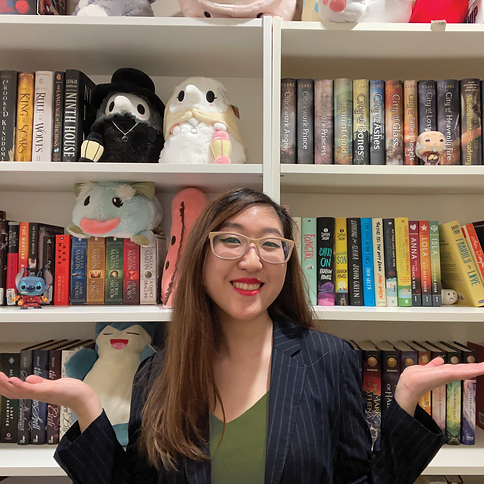 Girl with glasses, long brown hair, in front of a full bookshelf. She's wearing a black and white striped blazer and a green shirt.